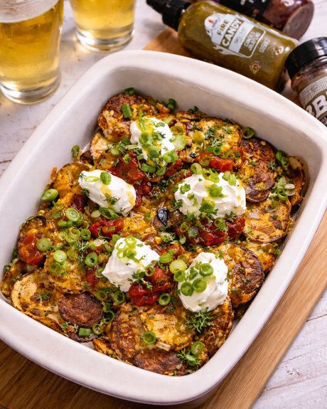 Crispy Irish nachos made with roasted potato slices topped with melted cheddar, bacon, salsa, sour cream, and sliced green onions, served in a white baking dish with Maritime Madness hot sauces and two glasses of beer in the background.