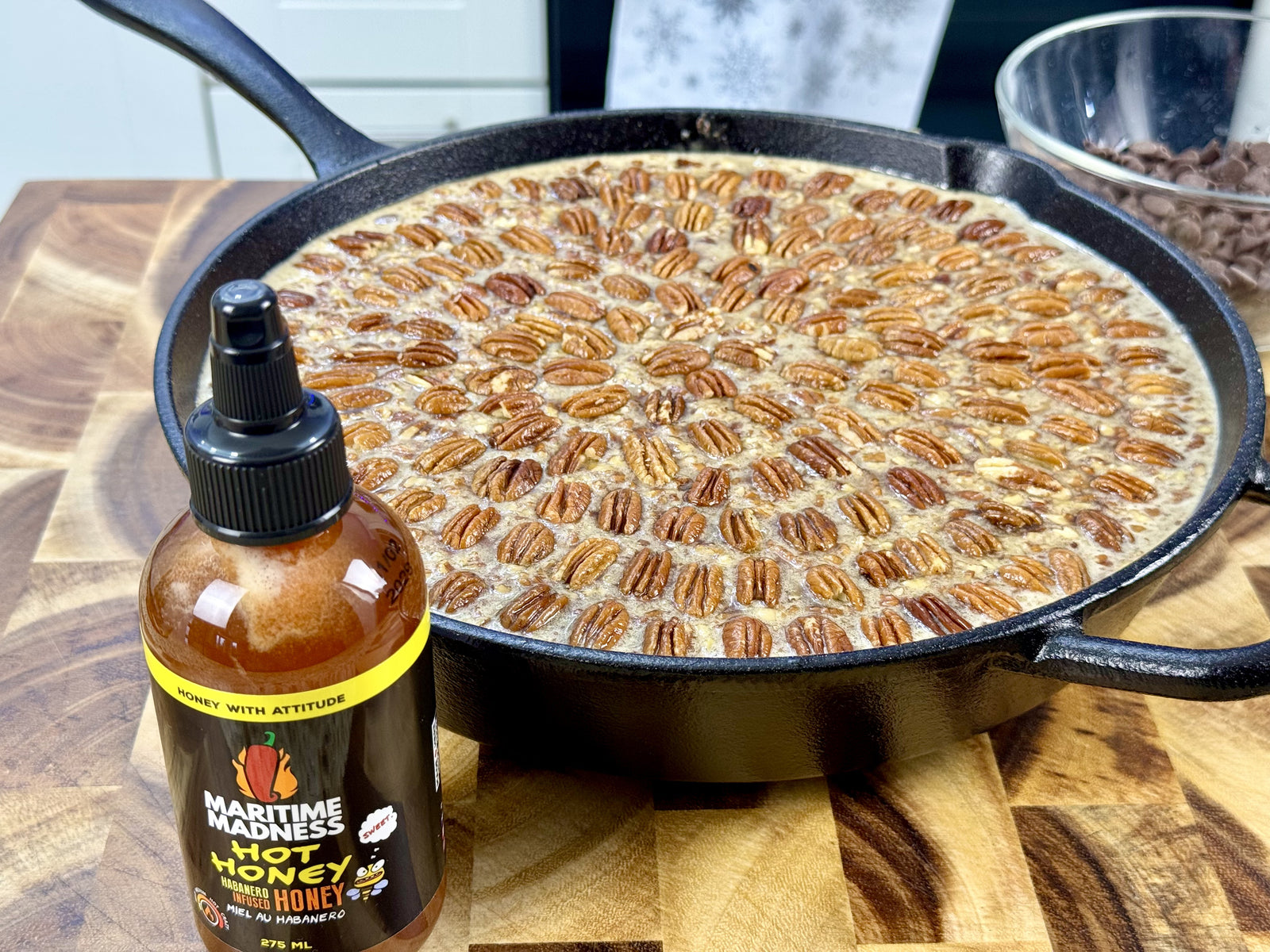 Maritime Madness Hot Honey beside a cast iron smoked pecan-chocolate cobbler topped with toasted pecans on a wooden cutting board.