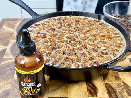 Maritime Madness Hot Honey beside a cast iron smoked pecan-chocolate cobbler topped with toasted pecans on a wooden cutting board.
