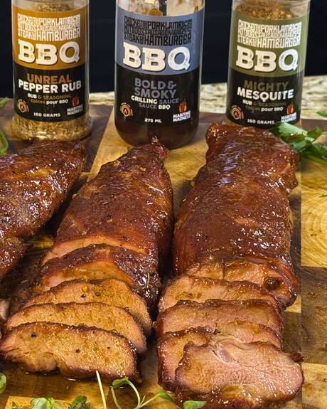 Three smoked pork tenderloins sliced on a wooden cutting board, glazed with a smoky BBQ sauce and cooked until juicy and caramelized. Maritime Madness Unreal Pepper Rub, Bold & Smokey Sauce, and Mighty Mesquite Rub are displayed behind the smoked pork.