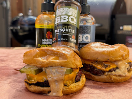 Smash burger topped with melted cheddar cheese, pickles, and creamy burger sauce dripping over the sides on a toasted bun. Maritime Madness Mighty Mesquite Rub and sauces are displayed behind the burger beside a smoker in the background.