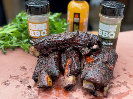 Close-up of smoked beef back ribs with a dark caramelized bark stacked on butcher paper, seasoned with Maritime Madness Mighty Mesquite and Unreal Pepper Rub. Bottles of Maritime Madness BBQ sauce and rubs sit behind the ribs beside fresh herbs.