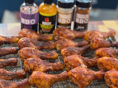 Smoked chicken drumsticks seasoned with BBQ rubs and arranged on cooling racks, with Maritime Madness sauces and seasonings displayed in the background. The drumsticks have a crispy, smoky exterior with a rich golden-brown colour from the smoker.