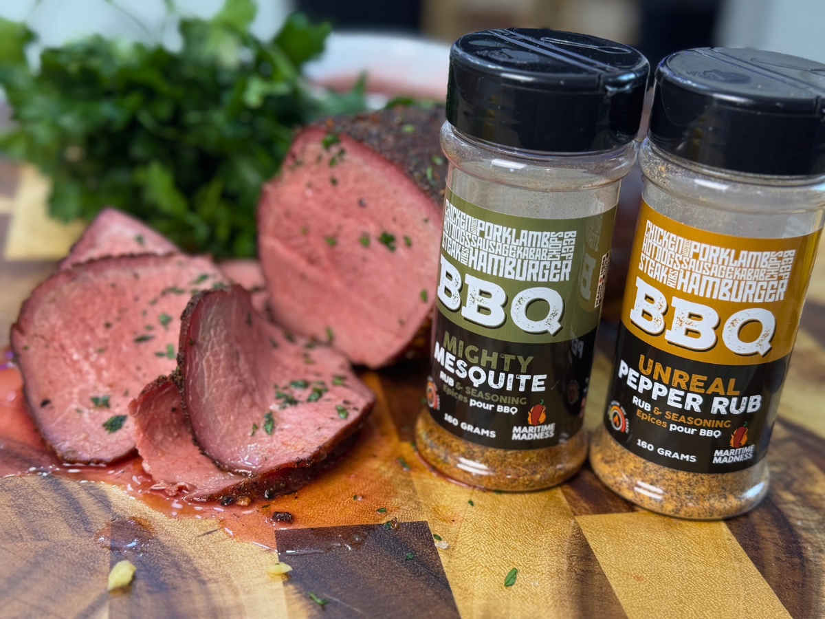 Sliced smoked beef eye round cooked medium-rare on a wooden cutting board beside Maritime Madness Mighty Mesquite and Unreal Pepper Rubs. The roast has a smoky crust with juicy pink slices sprinkled with fresh herbs.