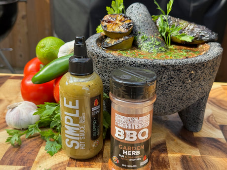 Bottle of Maritime Madness Simple Jalapeno & Lime hot sauce and Garden Herb BBQ rub on a cutting board beside fresh limes, jalapeno, tomato, garlic, herbs, and a stone mortar filled with charred salsa ingredients.