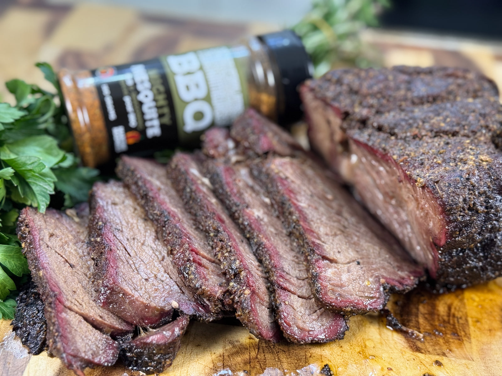 Sliced smoked chuck roast with a dark bark and pink smoke ring on a wooden cutting board, seasoned with Maritime Madness Mighty Mesquite rub and served with fresh herbs in the background.