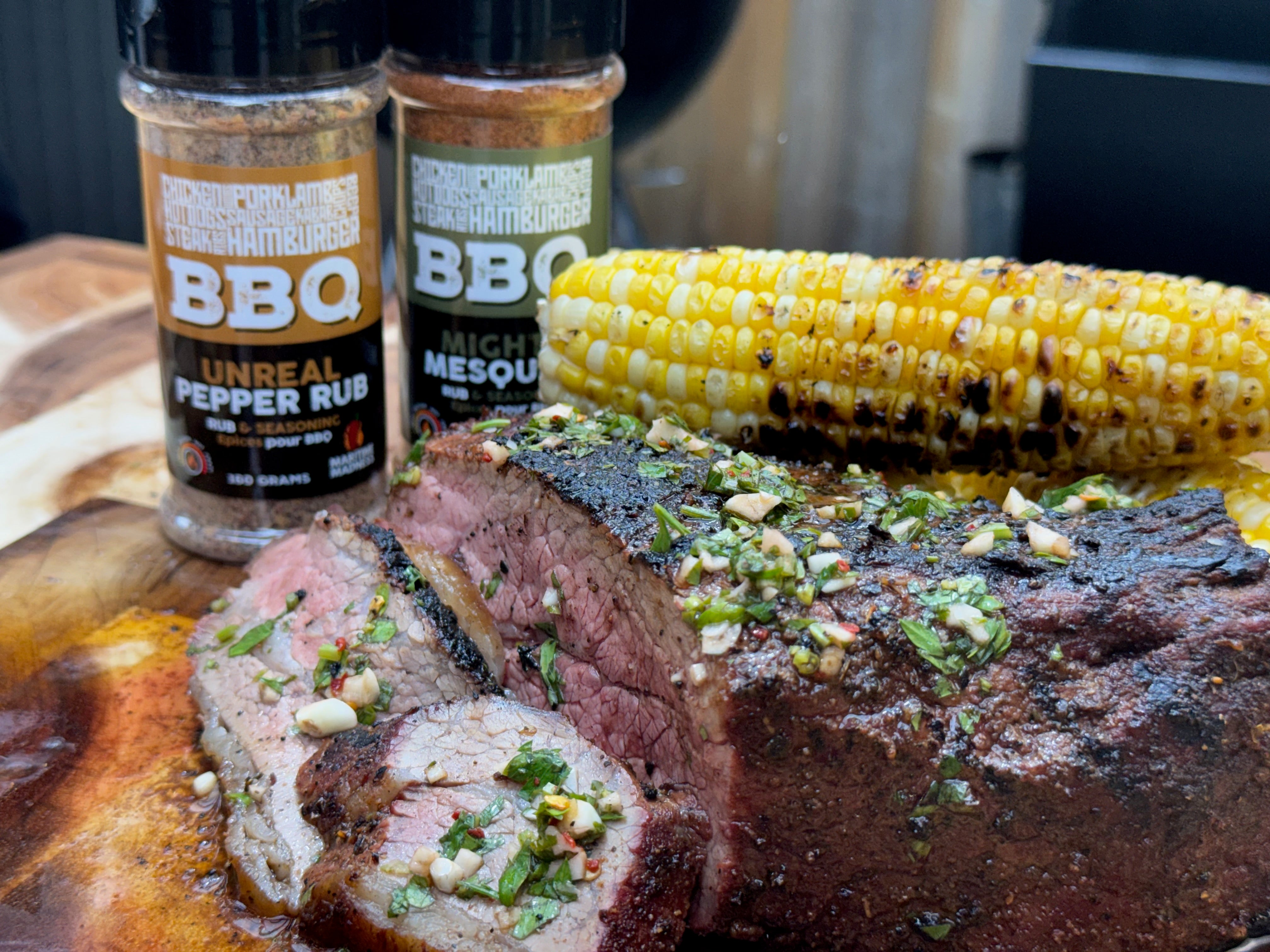 Sliced smoked tri tip topped with fresh chimichurri on a wooden cutting board, served with grilled corn and Maritime Madness Mighty Mesquite and Unreal Pepper BBQ rubs in the background.