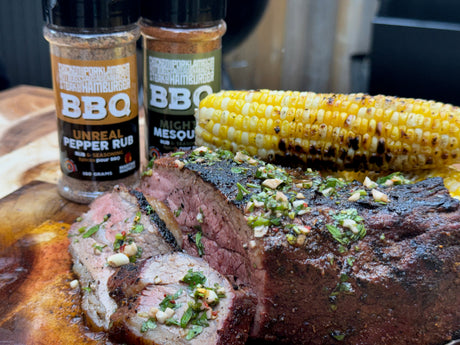 Sliced smoked tri tip topped with fresh chimichurri on a wooden cutting board, served with grilled corn and Maritime Madness Mighty Mesquite and Unreal Pepper BBQ rubs in the background.