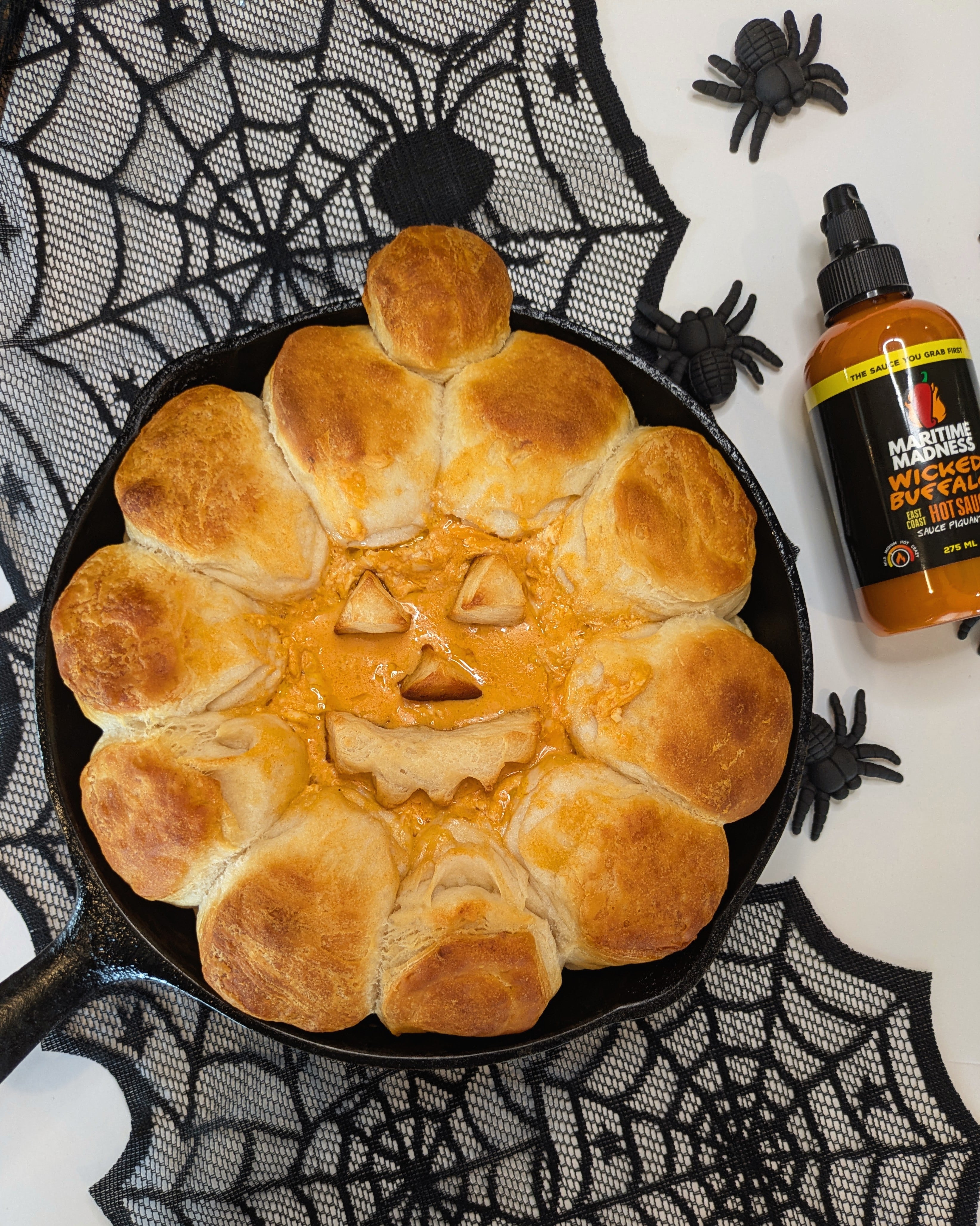 Jack O’Lantern Buffalo Chicken Dip baked in a cast iron skillet with golden rolls, spicy Wicked Buffalo chicken dip, Halloween spiderweb décor, and a bottle of Maritime Madness Wicked Buffalo Sauce.