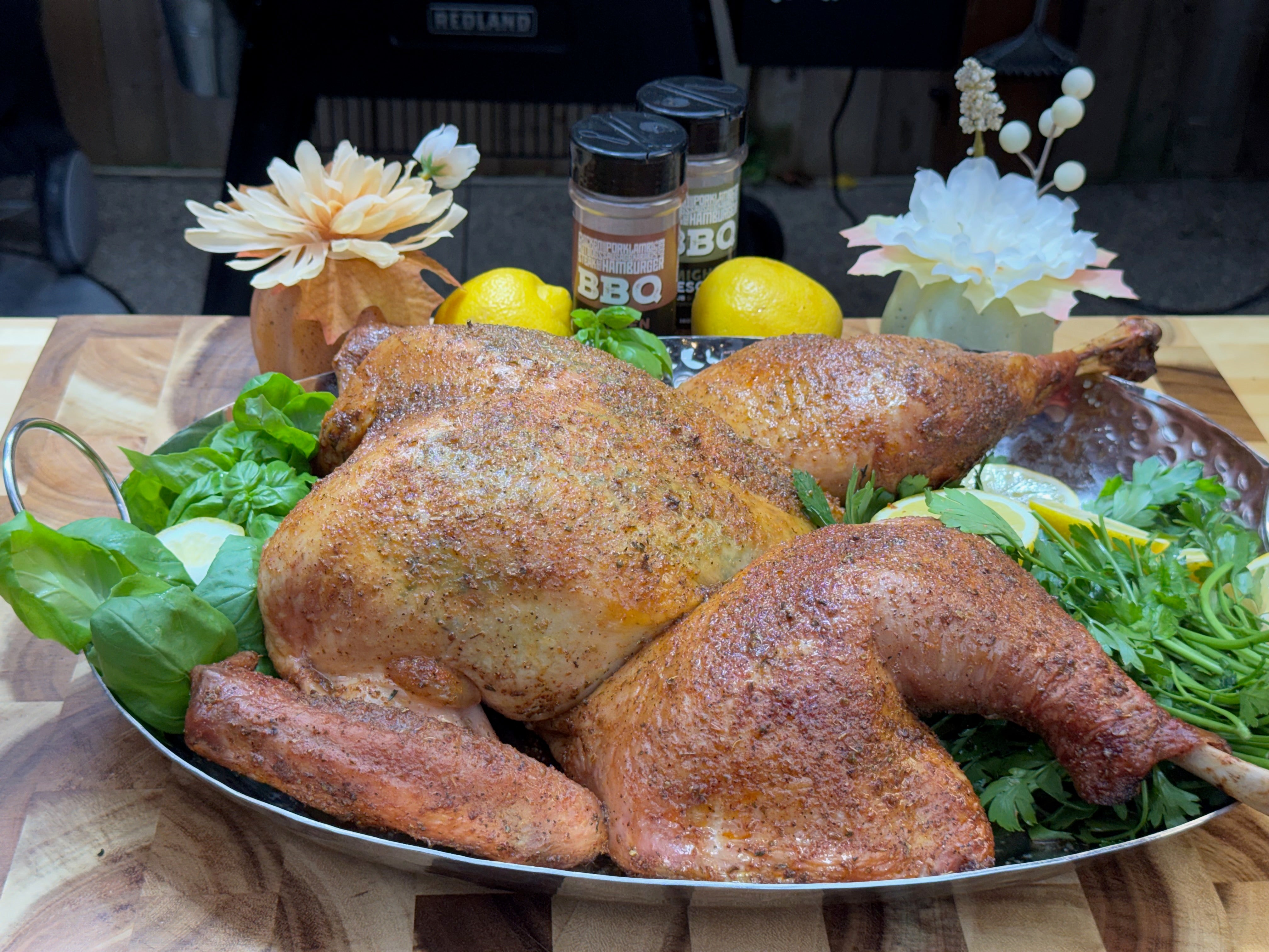 Golden-brown roasted turkey on a silver platter, surrounded by fresh herbs and lemon slices, with two bottles of BBQ seasoning and decorative flowers behind it.