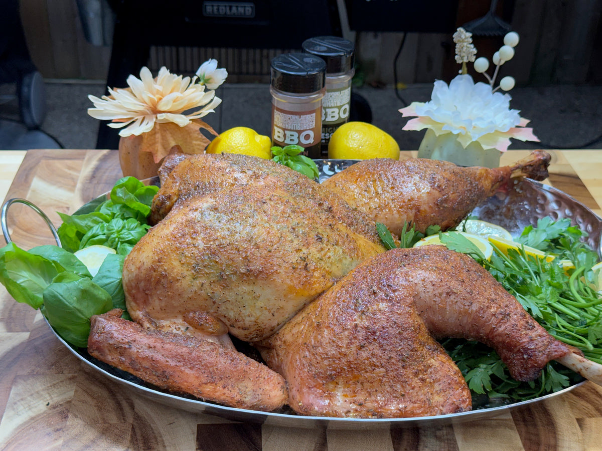 Golden-brown roasted turkey on a silver platter, surrounded by fresh herbs and lemon slices, with two bottles of BBQ seasoning and decorative flowers behind it.