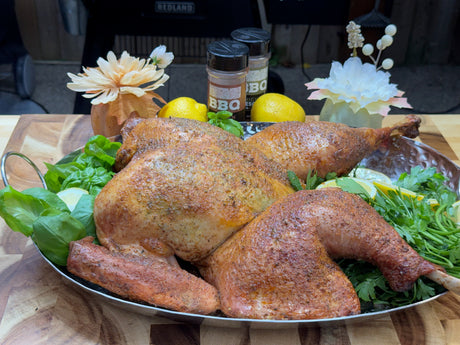 Golden-brown roasted turkey on a silver platter, surrounded by fresh herbs and lemon slices, with two bottles of BBQ seasoning and decorative flowers behind it.